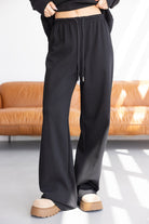 מכנסי טרנינג שחורים לנשים בגזרה רחבה עם גומי ומיתר, צילום בסטודיו עם ספה חומה ברקע