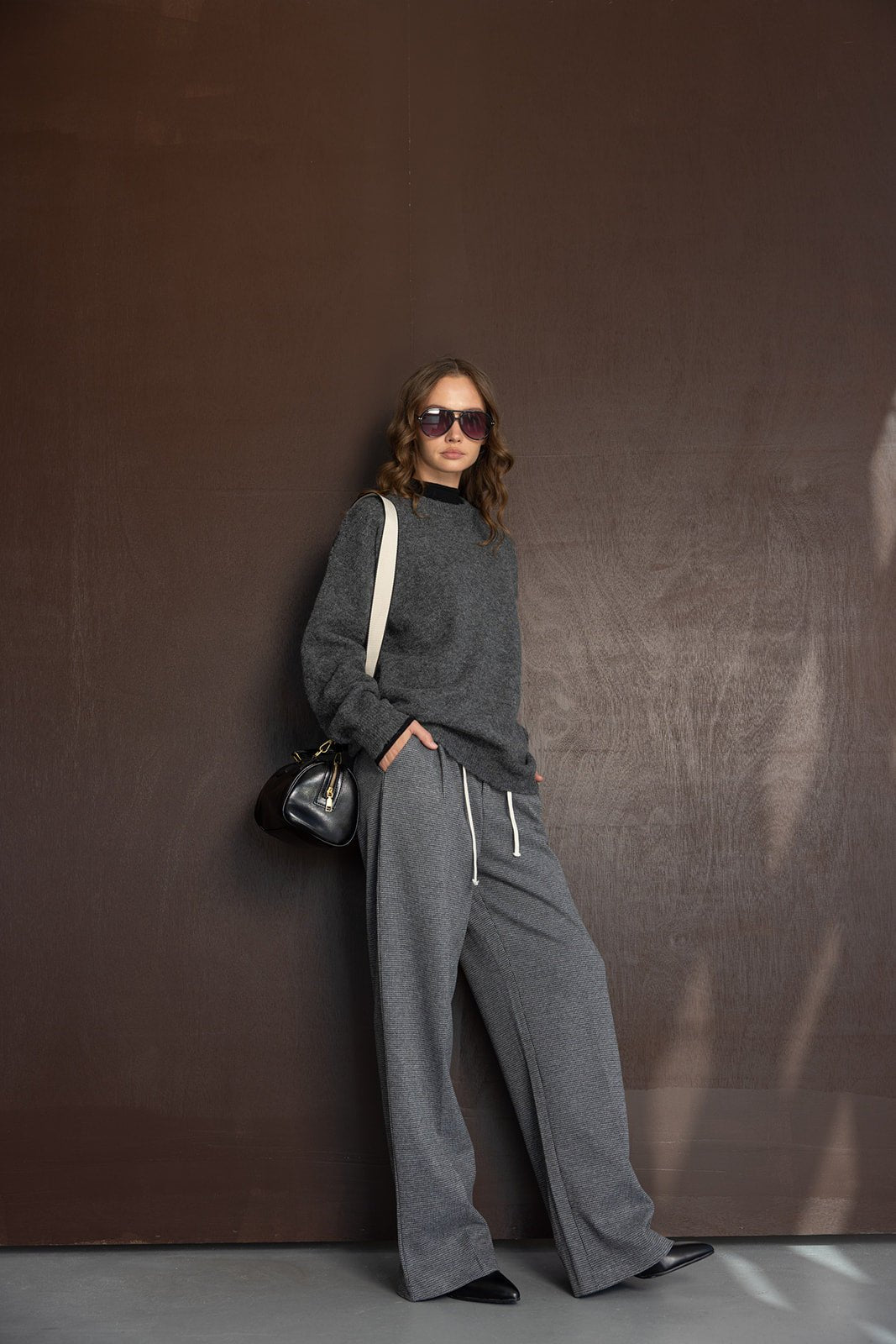 אישה לובשת בגדי אופנה לנשים, סווטשרט אפור ומכנסיים רחבים, משקפי שמש ותיק יד שחור