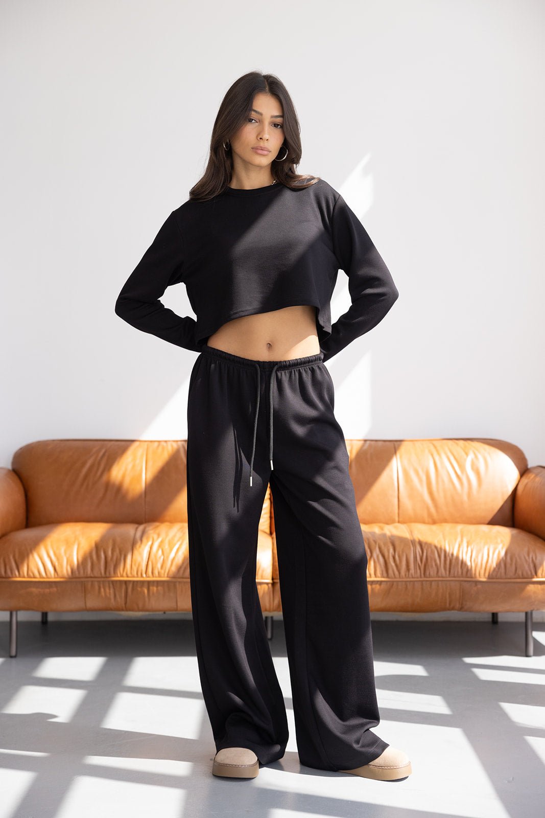 דוגמנית לובשת סט בגדי נשים שחור עם חולצה קרופ ארוכה ומכנסיים רחבים בסטייל קז'ואל, רקע סלון מודרני