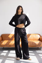 דוגמנית לובשת סט בגדי נשים שחור עם חולצה קרופ ארוכה ומכנסיים רחבים בסטייל קז'ואל, רקע סלון מודרני