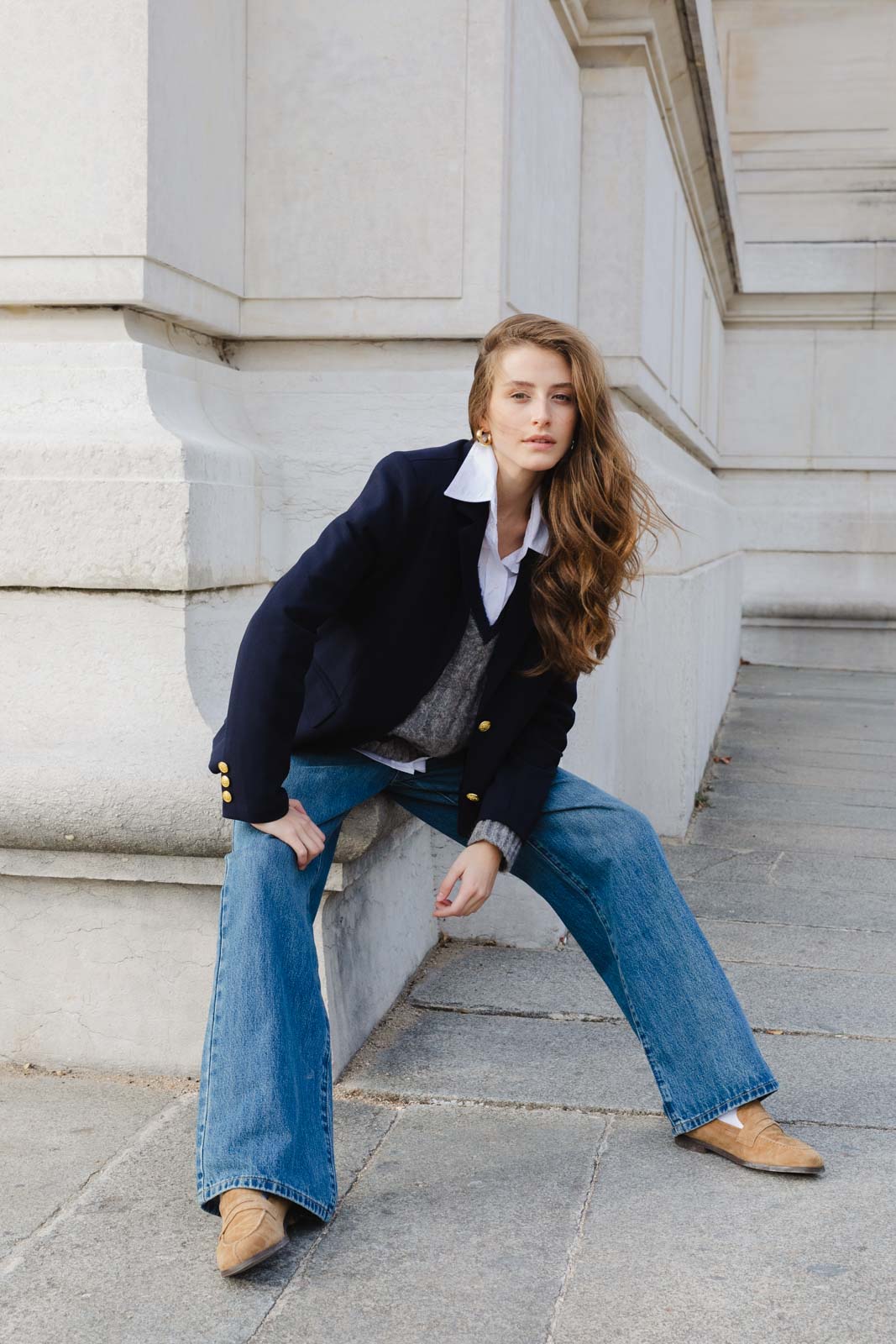 דוגמנית צעירה בלבוש אופנתי: בלייזר כחול, חולצה לבנה, ג'ינס רחב ונעליים חומות, יושבת ליד קיר אבן