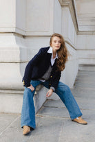 דוגמנית צעירה בלבוש אופנתי: בלייזר כחול, חולצה לבנה, ג'ינס רחב ונעליים חומות, יושבת ליד קיר אבן