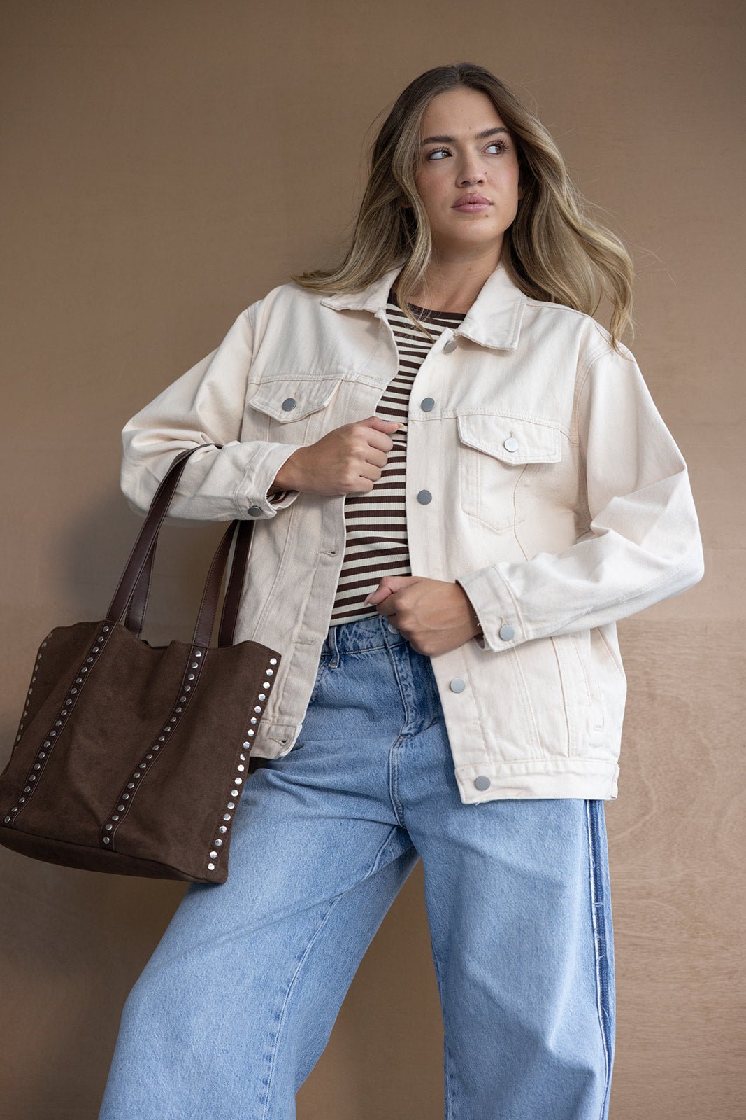 אישה עם ג'קט ג'ינס בז', חולצת פסים חומה ומכנסי ג'ינס רחבים, תיק חום אופנתי