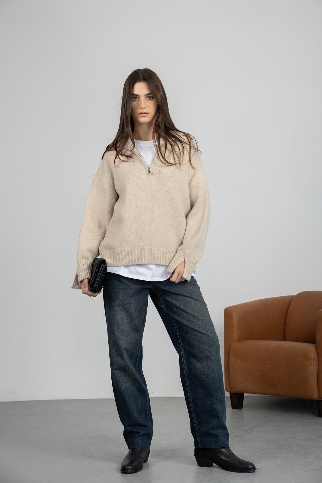 אישה לובשת סוודר בז' וג'ינס רחב, תיק קלאץ' שחור ונעלי עקב שחורות, אופנת נשים קז'ואל