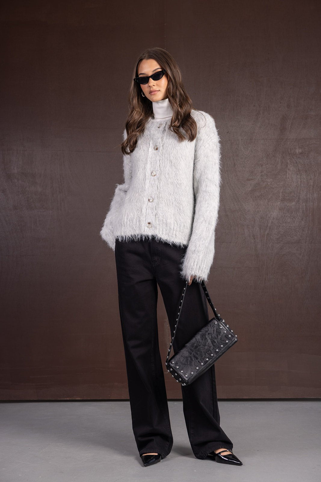 אישה עם קרדיגן אפור פרוותי, מכנסיים שחורים רחבים, משקפי שמש ותיק שחור אופנתי