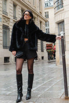 אישה אופנתית ברחוב עירוני לובשת מעיל פרווה שחור, מכנסיים קצרים, גרביון ומגפיים גבוהים