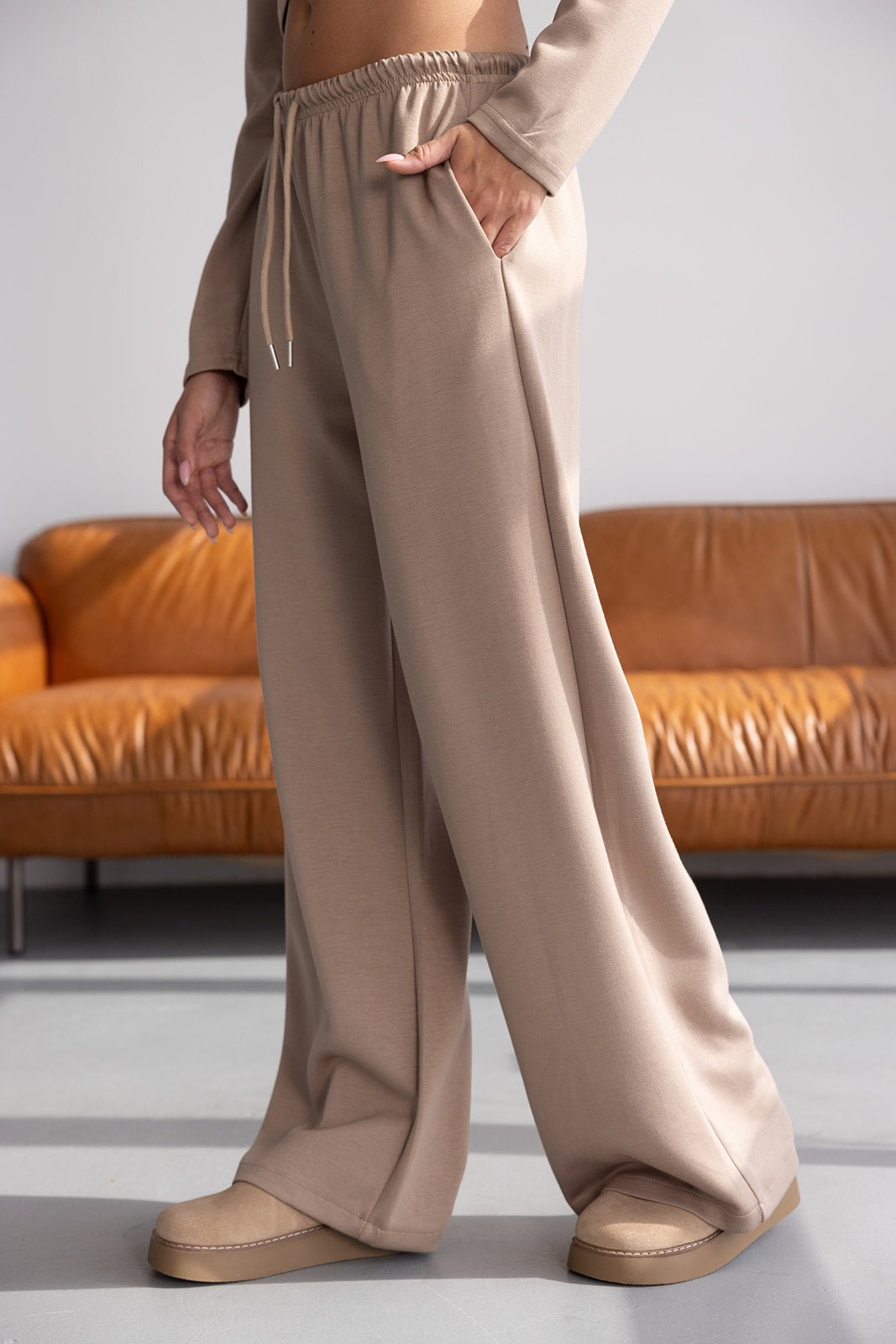 מכנסיים רחבים בצבע בז' לנשים, גזרה גבוהה, בד נוח, מצולמים ליד ספה חומה