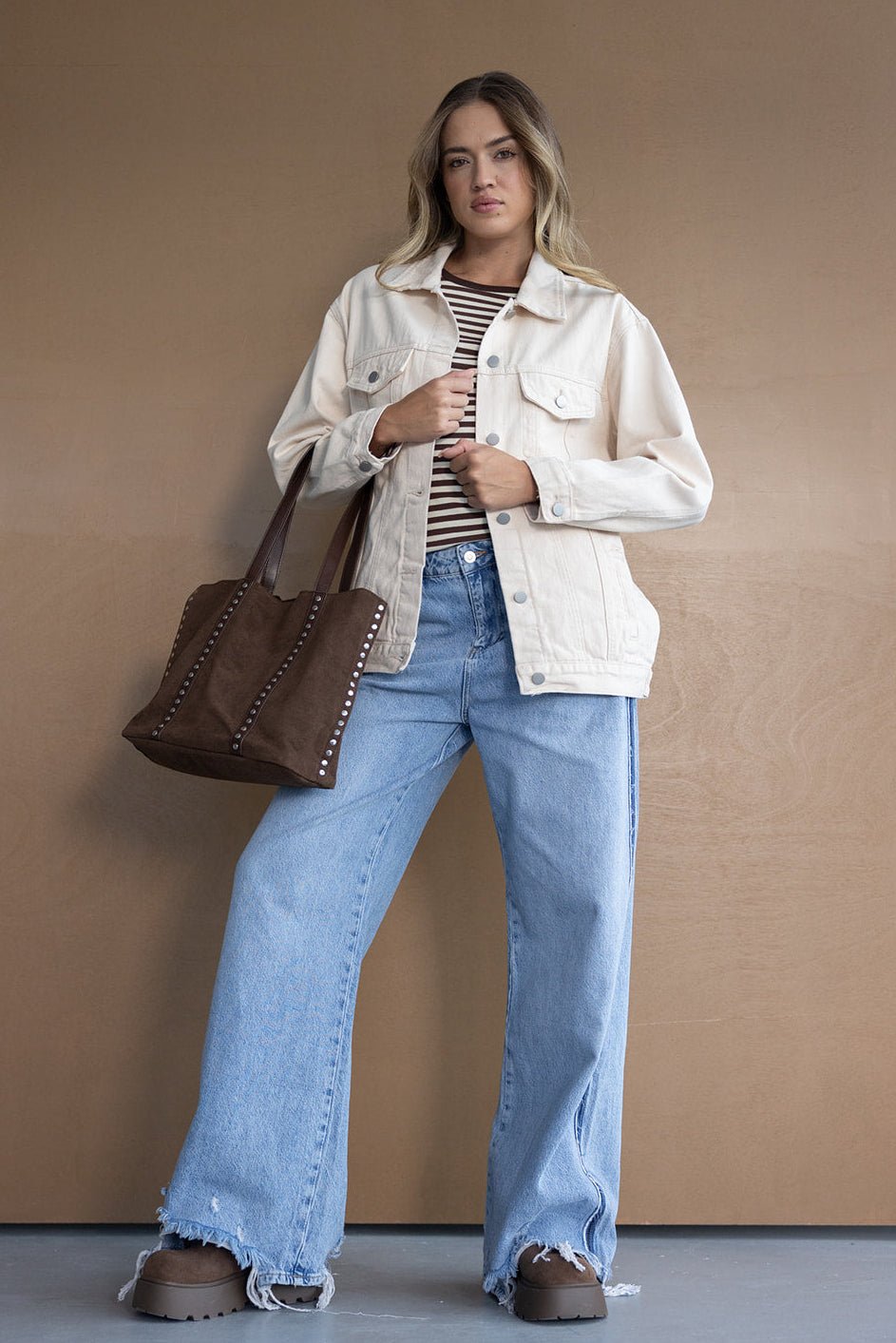 אישה צעירה עם ג'קט ג'ינס בהיר, חולצת פסים, ג'ינס בגזרה רחבה ותיק חום, לוק אופנתי לנשים