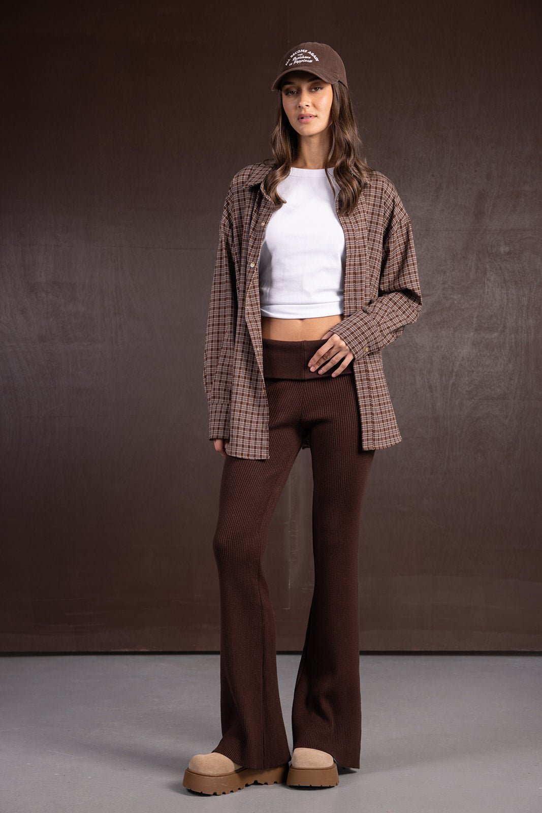 אישה לובשת מכנסיים חומים מתרחבים, גופיה לבנה קצרה, חולצה משובצת וכובע חום אופנתי