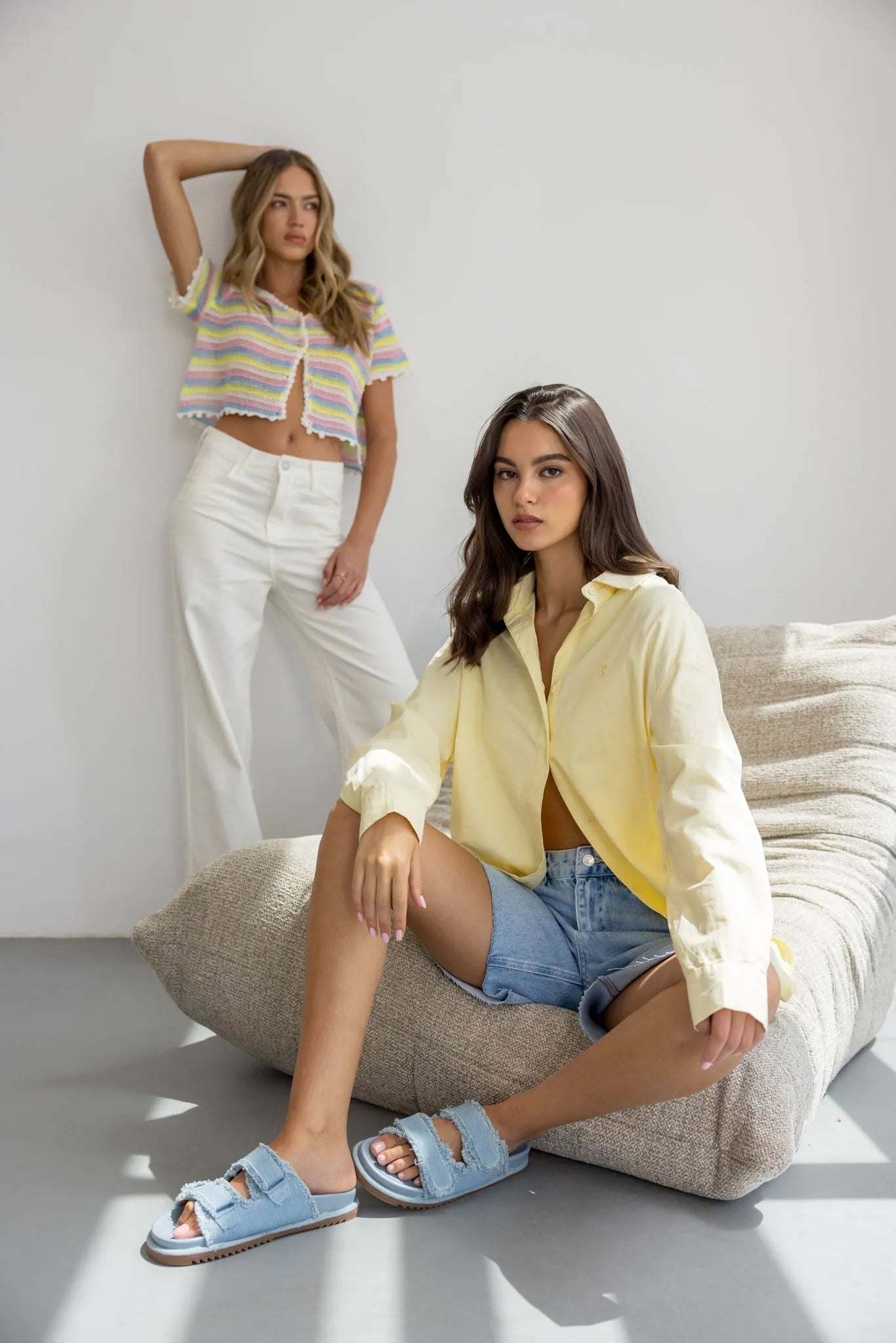 שתי נשים מצולמות בסגנון אופנתי, אחת יושבת עם חולצה צהובה, מכנסי ג’ינס וסנדלים כחולים, השנייה עומדת עם מכנסיים לבנים וחולצה צבעונית קייצית