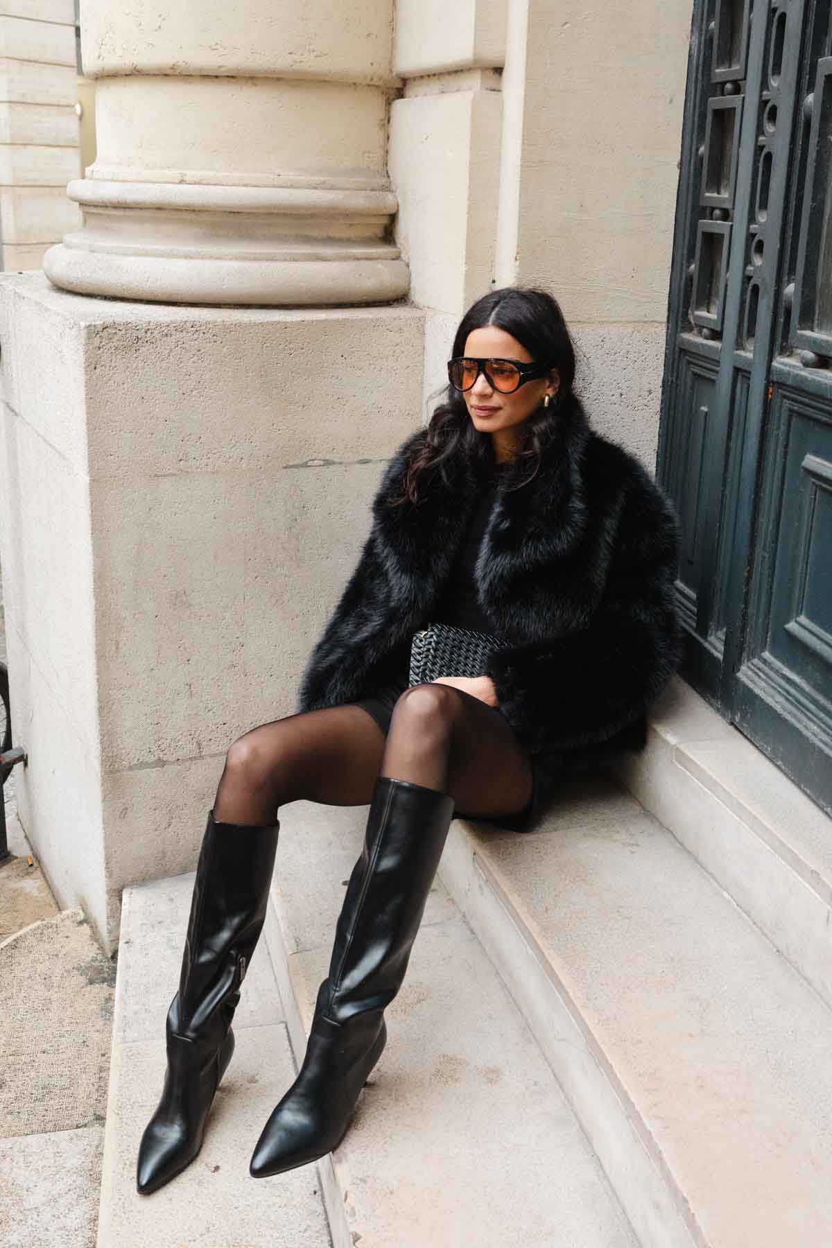 אישה לובשת מעיל פרווה שחור, גרביונים ומגפיים גבוהים אופנתיים, יושבת על מדרגות בעיר