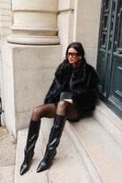 אישה לובשת מעיל פרווה שחור, גרביונים ומגפיים גבוהים אופנתיים, יושבת על מדרגות בעיר