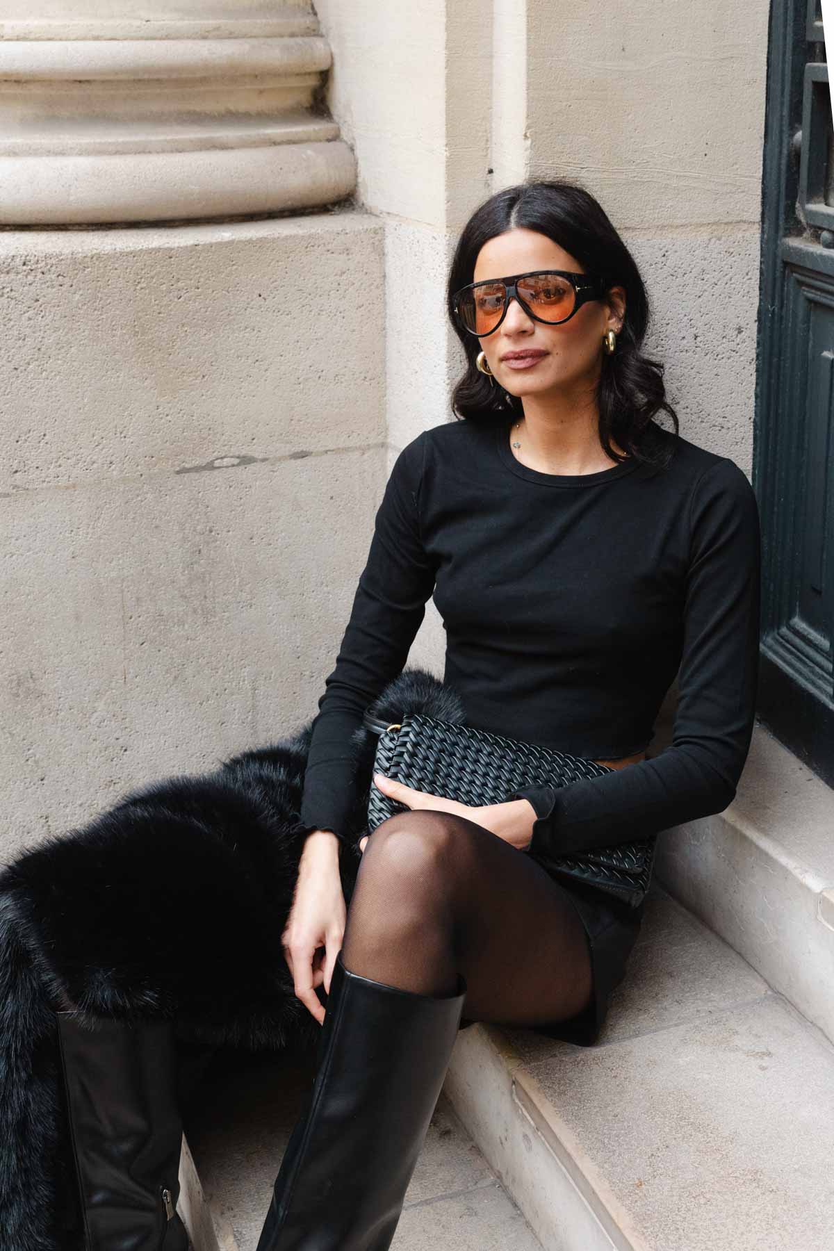 אישה יושבת בלבוש אופנתי, חולצה שחורה, חצאית קצרה, מגפיים גבוהים ותיק ערב, לוק אורבני לנשים