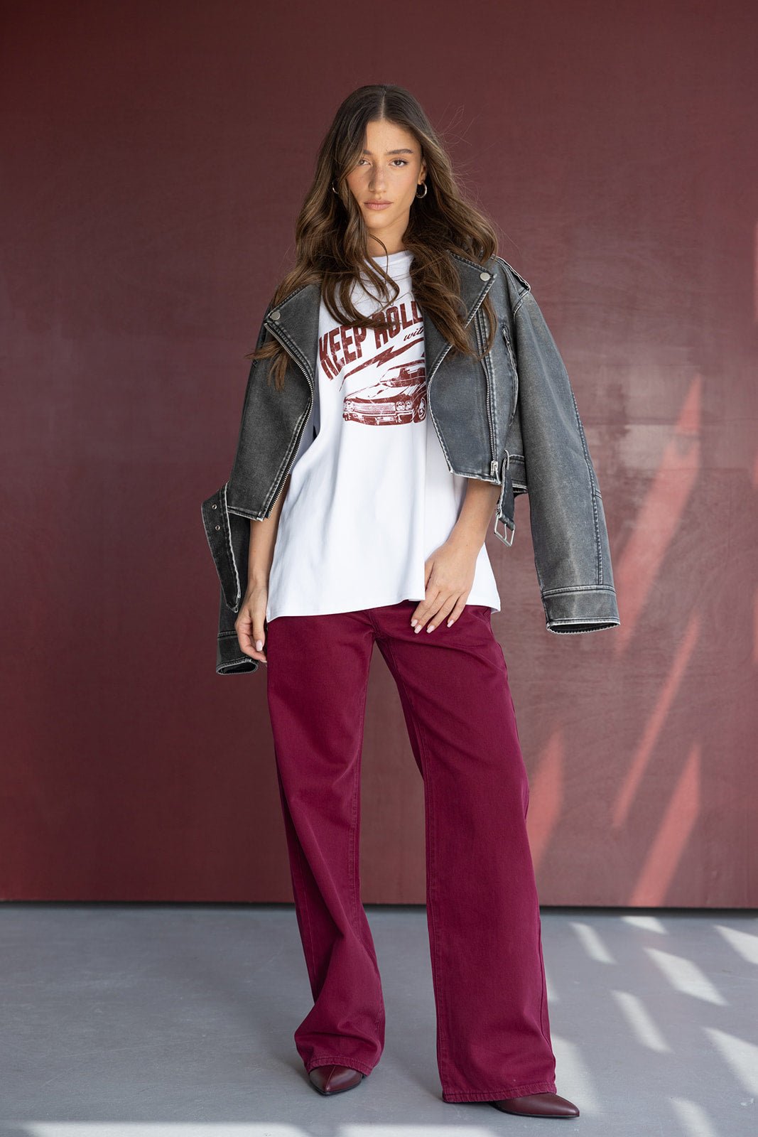 דוגמנית עם מכנסיים רחבים בורדו, ג'קט ג'ינס שחור וחולצת הדפס אופנתית, עומדת על רקע אדום