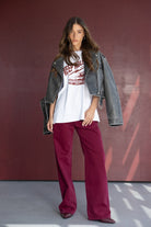 דוגמנית עם מכנסיים רחבים בורדו, ג'קט ג'ינס שחור וחולצת הדפס אופנתית, עומדת על רקע אדום