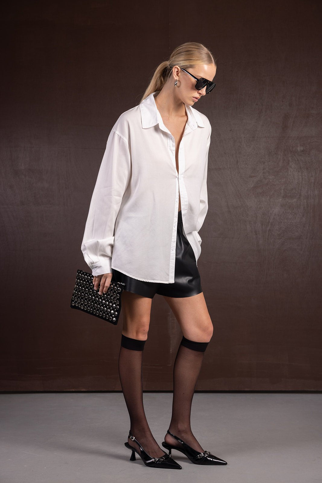 אישה בלוק אופנתי עם חולצה לבנה רחבה, חצאית מיני דמוי עור שחורה, גרבונים שחורים, נעלי עקב ותיק קלאץ' שחור עם ניטים.