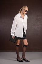 אישה בלוק אופנתי עם חולצה לבנה רחבה, חצאית מיני דמוי עור שחורה, גרבונים שחורים, נעלי עקב ותיק קלאץ' שחור עם ניטים.