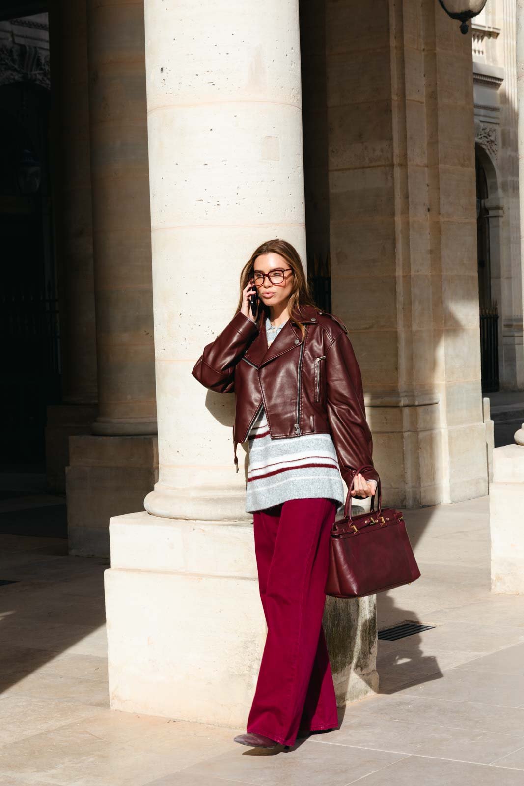 אישה בבגדי אופנה, מעיל עור חום, מכנסיים רחבים ובגד סריג, מחזיקה תיק ומתהלכת ברחוב עירוני