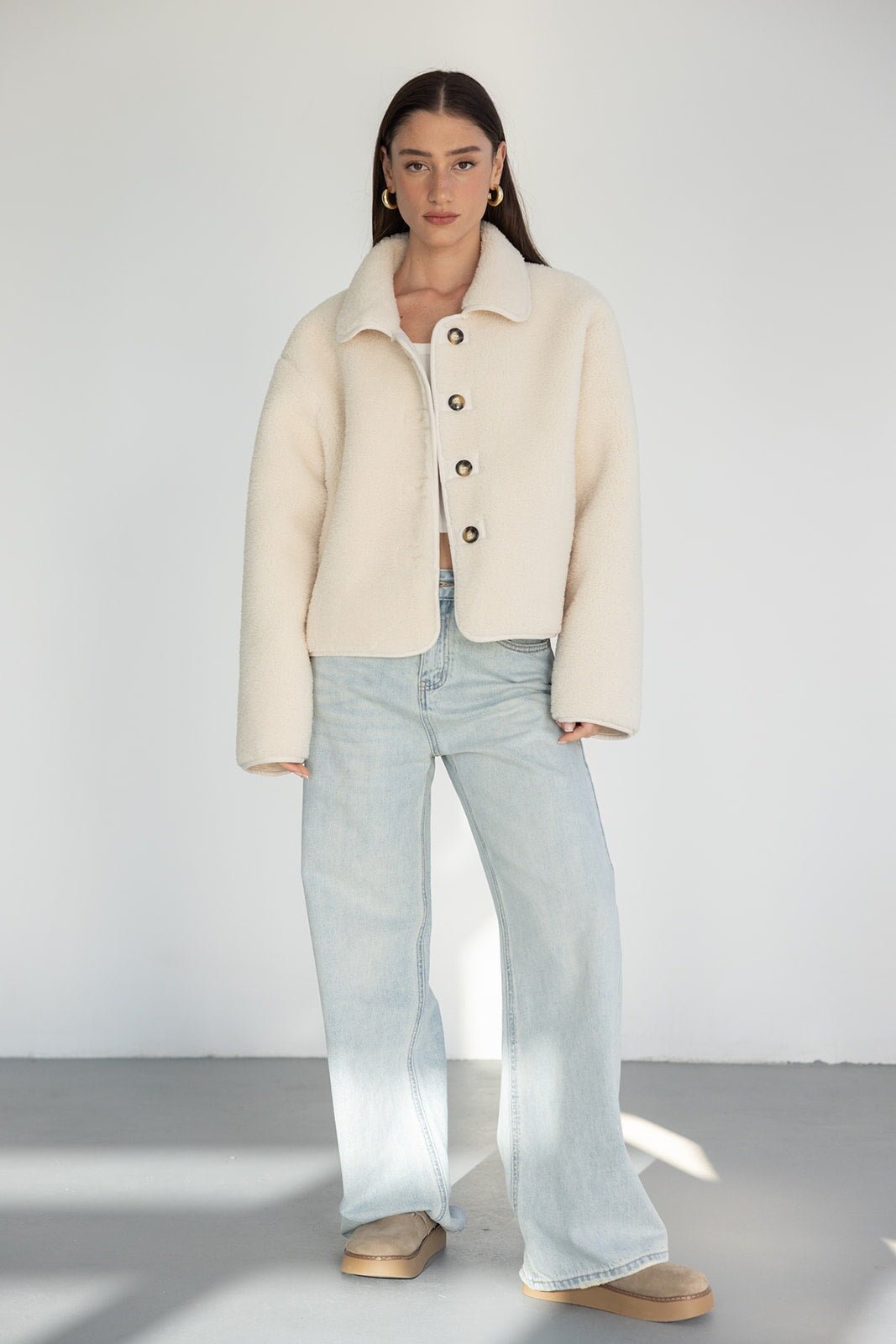 אישה לובשת מעיל צמר בז' עם כפתורים, ג'ינס רחב ובז' נעלי חורף אופנתיות, על רקע לבן