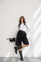 אישה עם חולצה לבנה, מכנסי ברמודה שחורים, מגפי בוקרים, תיק שחור ומשקפי שמש אופנתיים