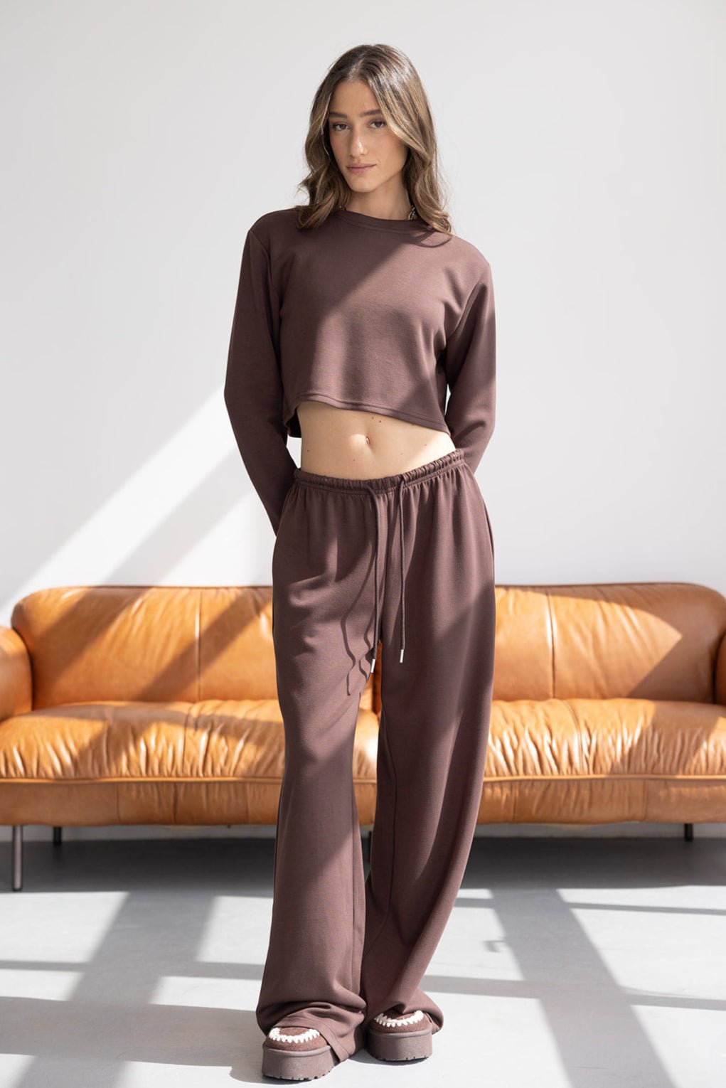 אשה צעירה בלבוש קז'ואל חום, חולצה קרופ ומכנס רחב, עומדת בסלון מודרני מואר