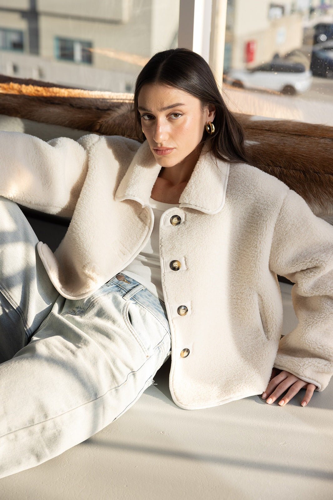 אישה צעירה עם שיער חום, לובשת מעיל פרוותי שמנת, טופ לבן וג'ינס אופנתי, יושבת באור טבעי