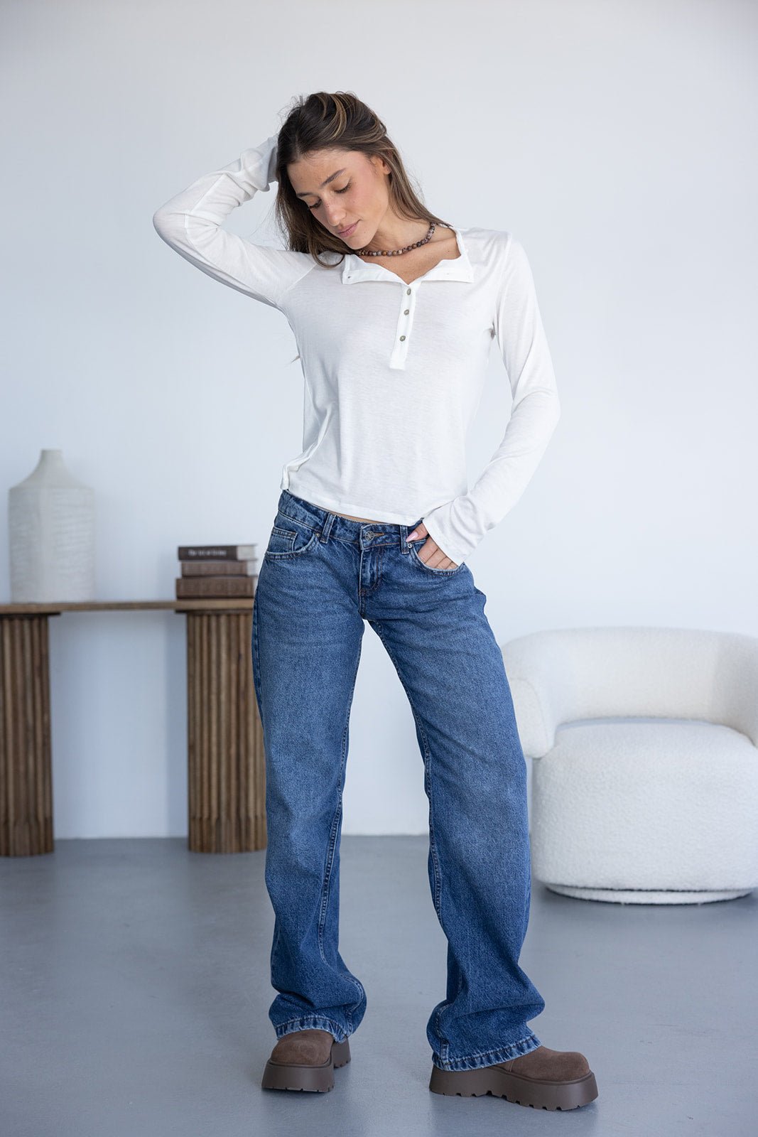 צעירה בלבוש קז'ואל לובשת ג'ינס כחול רחב, חולצת שרוול ארוך לבנה ונעלי פלטפורמה חומות, עומדת בחדר מודרני