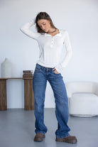 צעירה בלבוש קז'ואל לובשת ג'ינס כחול רחב, חולצת שרוול ארוך לבנה ונעלי פלטפורמה חומות, עומדת בחדר מודרני