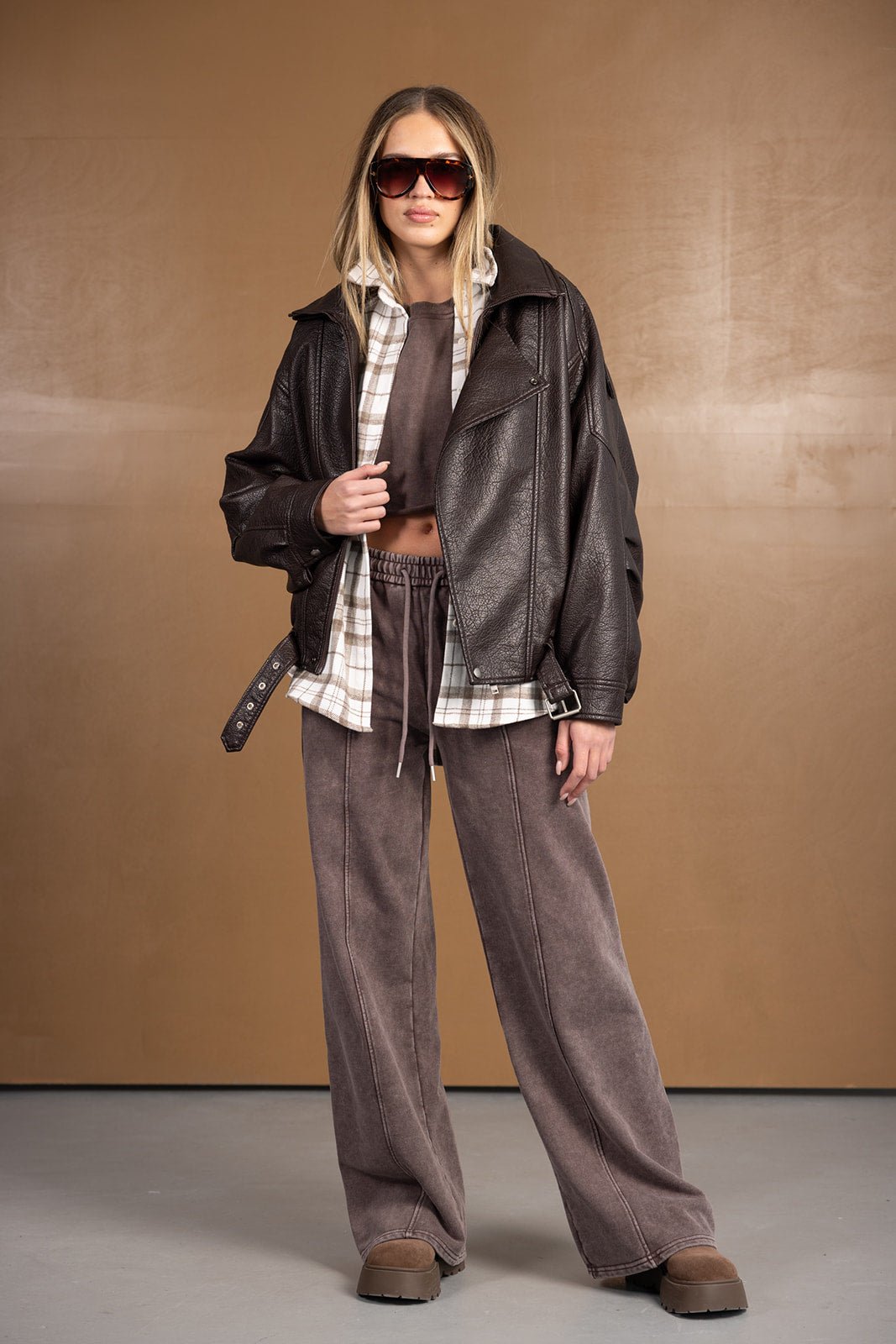 אישה עם משקפי שמש, לובשת ג'קט עור חום, חולצה משובצת ומכנסיים רחבים בגווני חום – אופנת חורף לנשים