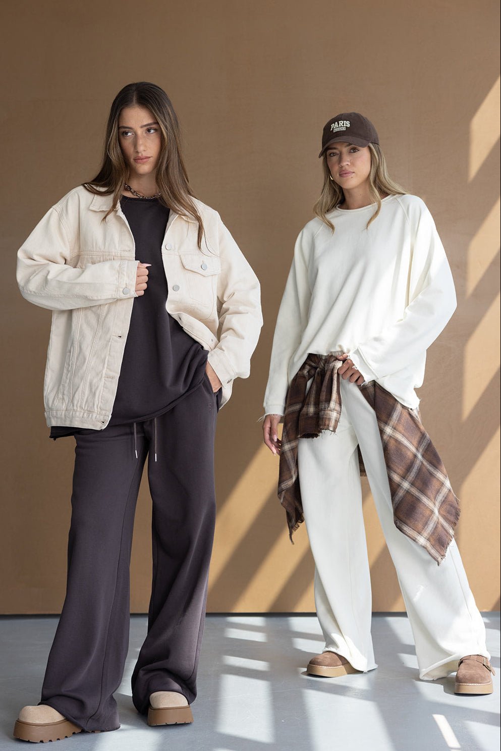 שתי נשים לובשות בגדי קז'ואל אופנתיים: אחת בחליפת טרנינג שחורה וז'קט בז', השנייה במכנס לבן, סווטשירט לבן וכובע, עם חולצה משובצת חומה קשורה למותן, רקע חום.