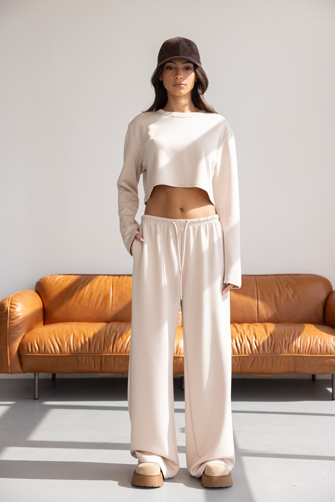 דוגמנית עם סט טרנינג בגוון בז', מכנסיים רחבים וחולצה קרופ, כובע מצחייה, יושבת על ספה חומה בסגנון מודרני