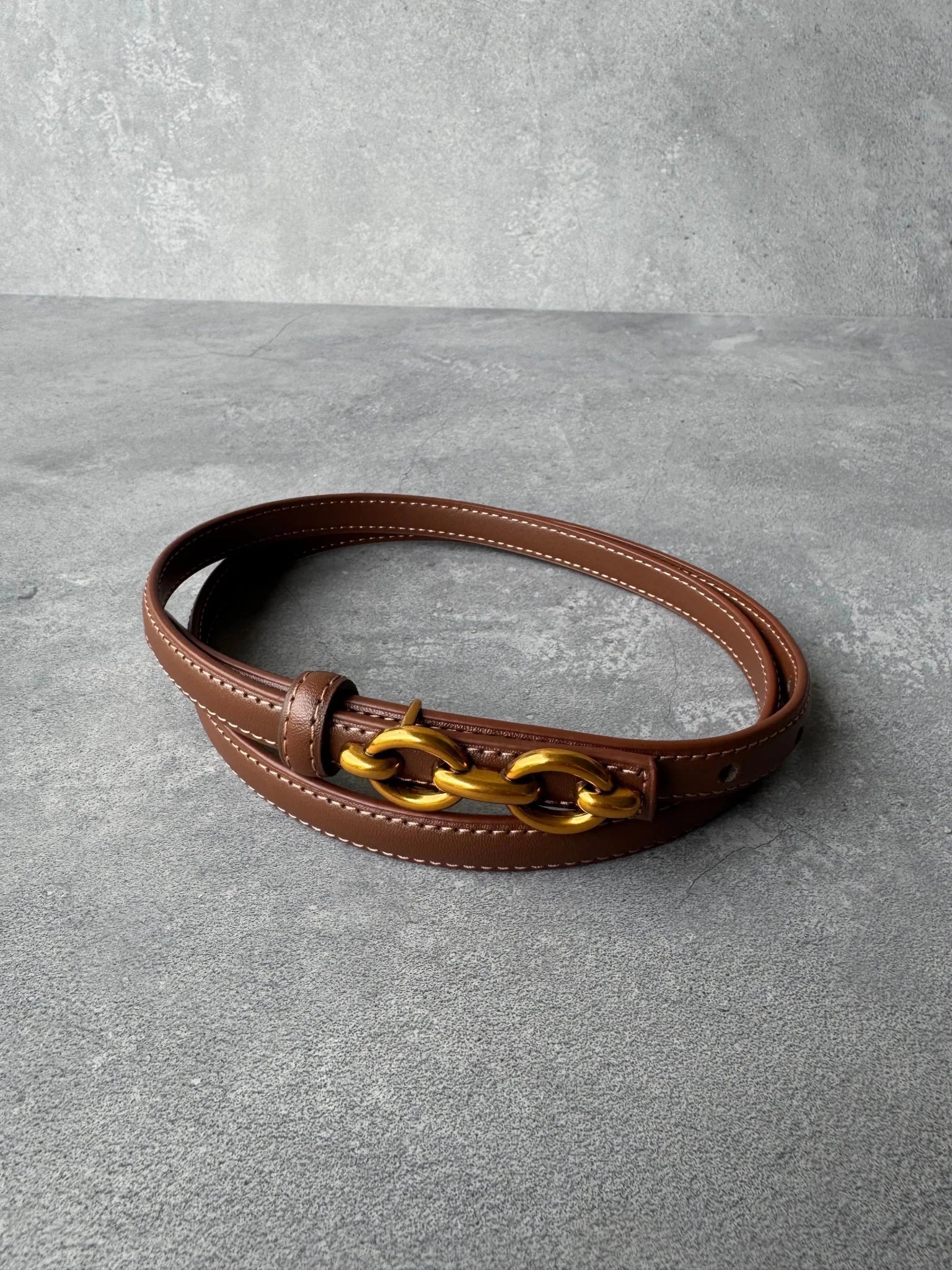 חגורת עור חומה עם אבזם שרשרת זהוב, אופנה לנשים ונערות, מונחת על משטח בטון