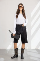 אישה עם חולצה לבנה ארוכה, מכנסיים קצרים שחורים, מגפיים גבוהים, תיק יד קלוע ומשקפי שמש אופנתיים