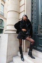 אופנת רחוב לנשים, דוגמנית בלבוש שחור עם ז׳קט פרווה, חצאית קצוצה, מגפיים גבוהים, מראה חורפי טרנדי בעיר אירופאית