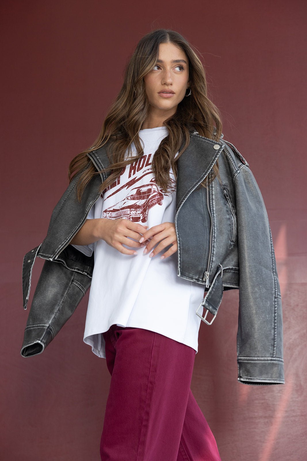 דוגמנית לובשת ג'קט ג'ינס אפור גדול, חולצה לבנה מודפסת ומכנסיים בורדו אופנתיים על רקע בורדו