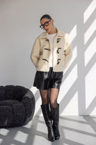 אישה עם משקפיים לובשת מעיל צמר לבן, מכנסי עור שחורים ומגפיים גבוהים לאופנת חורף