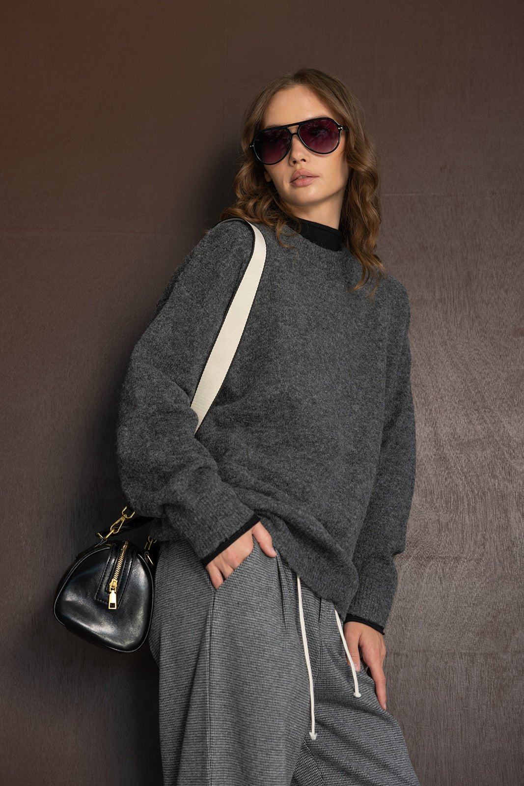 אישה לובשת סווטשירט אפור, מכנסיים אפורים ומשקפי שמש אופנתיים, עם תיק צד שחור