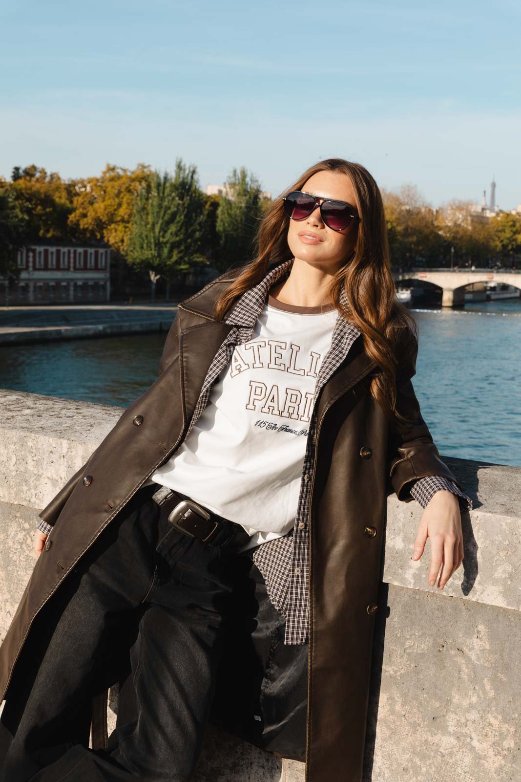 אישה אופנתית על רקע נהר, לובשת מעיל עור, משקפי שמש וחולצת ATELIER PARIS, סתיו עירוני