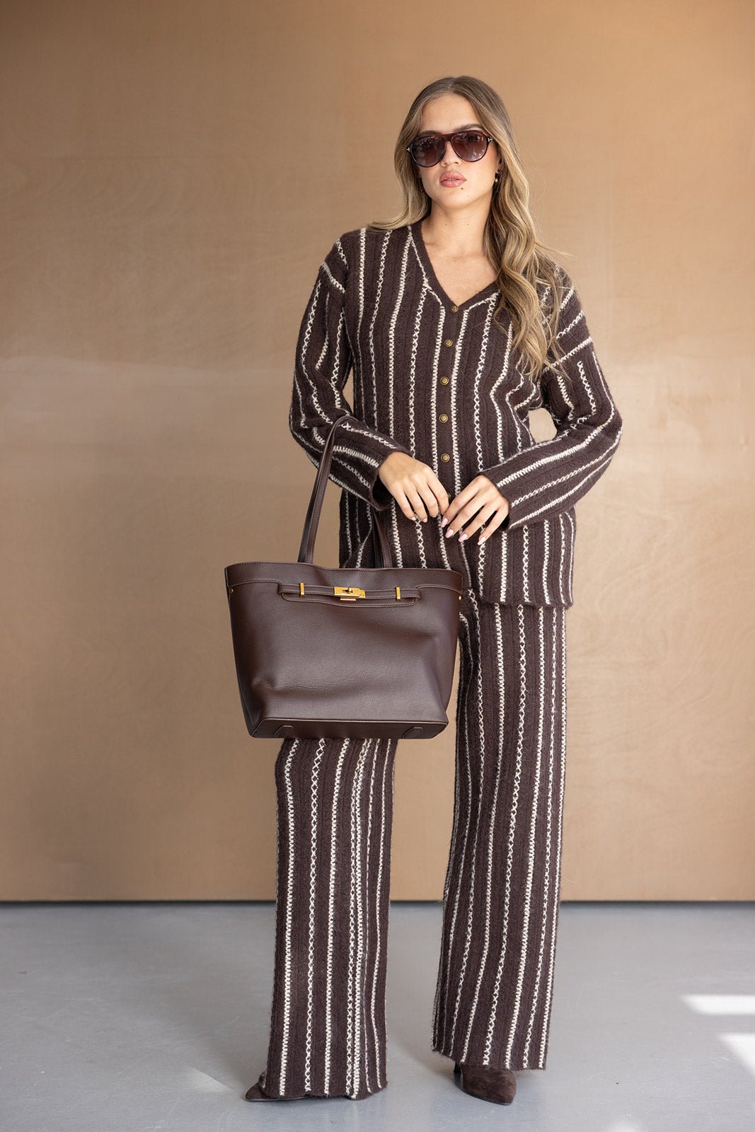 אישה לובשת סט פסים חום עם מכנסיים רחבים, סוודר תואם, תיק עור חום ומשקפי שמש אופנתיים