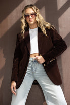 אישה עם בלייזר חום, ג'ינס בהיר, חולצה לבנה קצרה ומשקפי שמש אופנתיים, מראה רחוב טרנדי לנשים