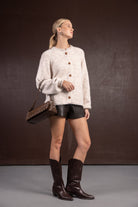 דוגמנית לובשת סוודר בז' עם כפתורים, מכנסי עור קצרים, מגפיים חומים ותיק צד חום