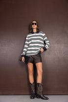 אישה עם חולצת פסים ארוכה, מכנסי עור קצרים, מגפיים שחורים ומשקפי שמש, לוק אופנתי לנשים