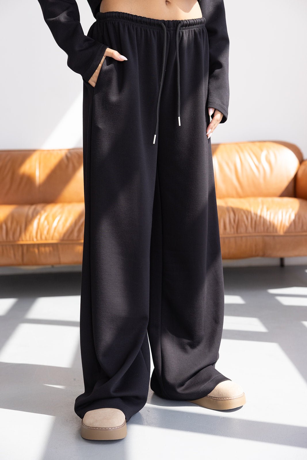 מכנסי נשים רחבים שחורים עם גומי ושרוך, נראים נוחים ובסגנון קז'ואל, רקע ספה חומה