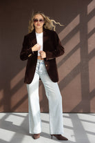 אישה עם ז'קט קטיפה חום, חולצה לבנה, ג'ינס רחב ומשקפי שמש אופנתיים