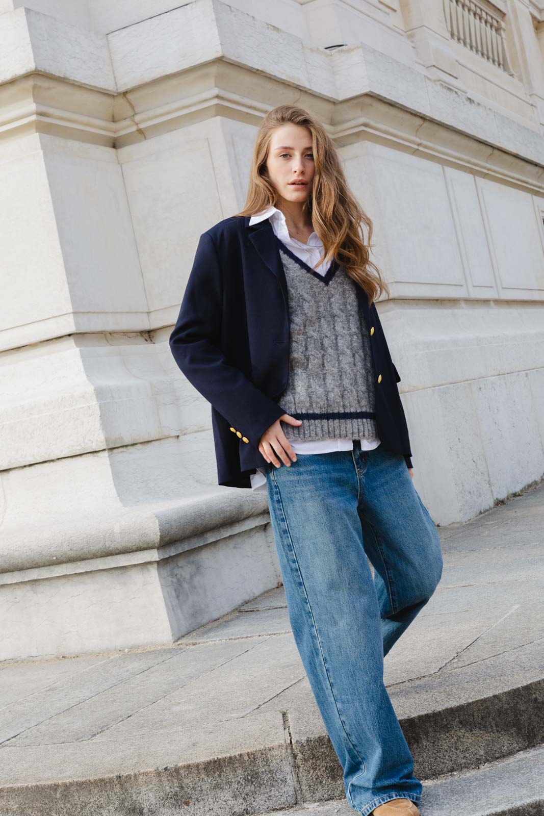 צעירה עם ג'ינס רחב, סוודר אפור, חולצה לבנה וז'קט כחול עומדת בסגנון אופנתי ליד קיר אבן