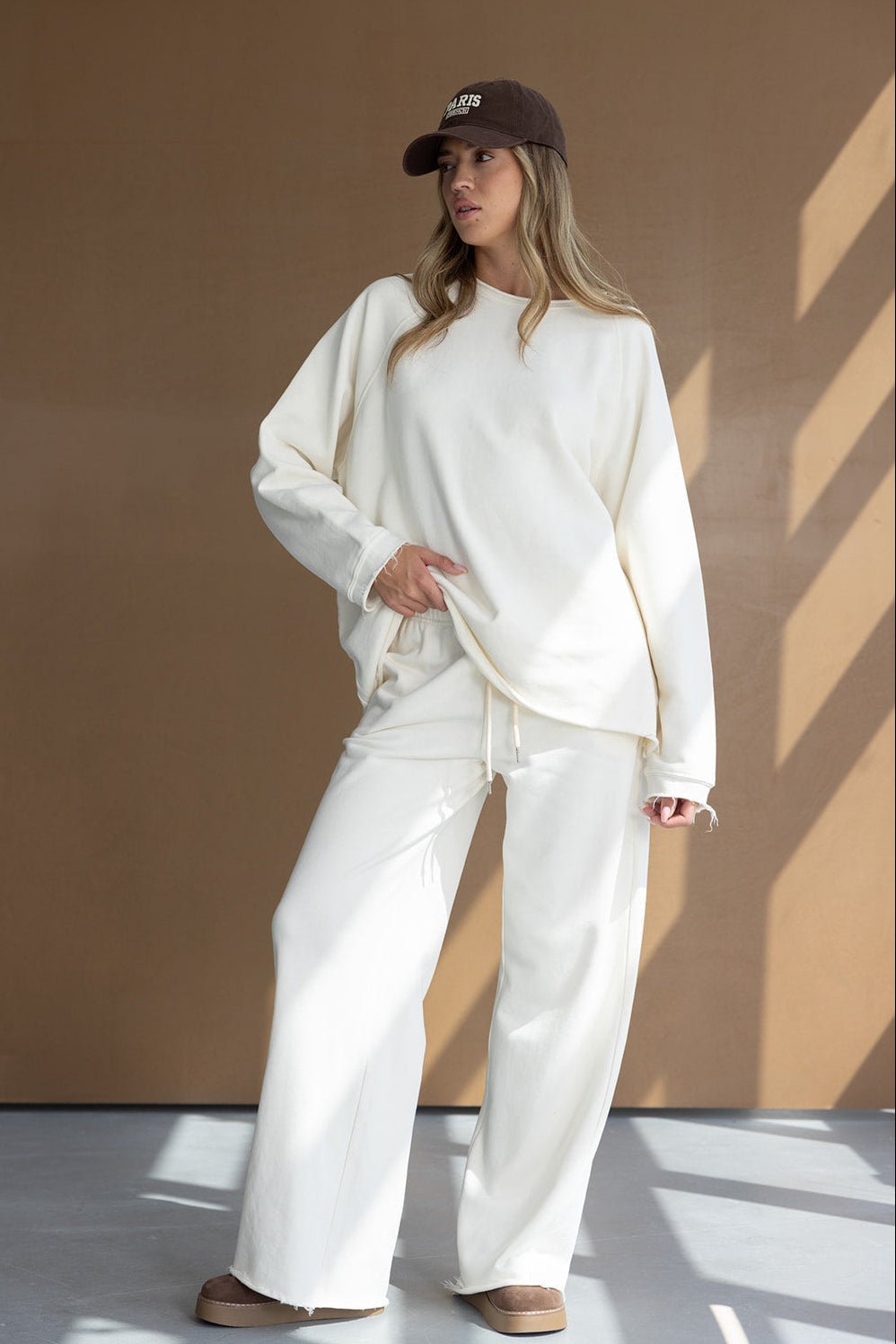 אישה לובשת סט טרנינג לבן רחב, כובע חום ונעלי בית אופנתיות, רקע חום מינימליסטי