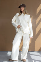 אישה לובשת סט טרנינג לבן רחב, כובע חום ונעלי בית אופנתיות, רקע חום מינימליסטי