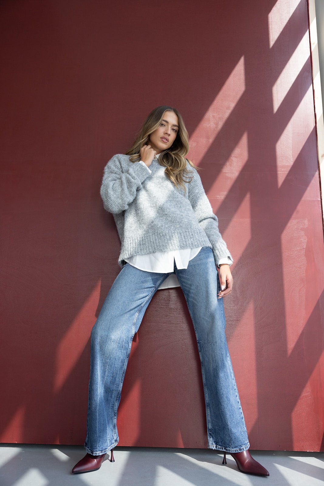 דוגמנית לובשת סוודר אפור ומכנסי ג'ינס רחבים, נעלי עקב בורדו, על רקע קיר אדום