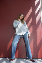 דוגמנית לובשת סוודר אפור ומכנסי ג'ינס רחבים, נעלי עקב בורדו, על רקע קיר אדום