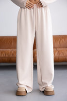 מכנסי טרנינג רחבים בצבע בז' לנשים, גומי במותן, נעלי פלטפורמה, רקע סלון חום